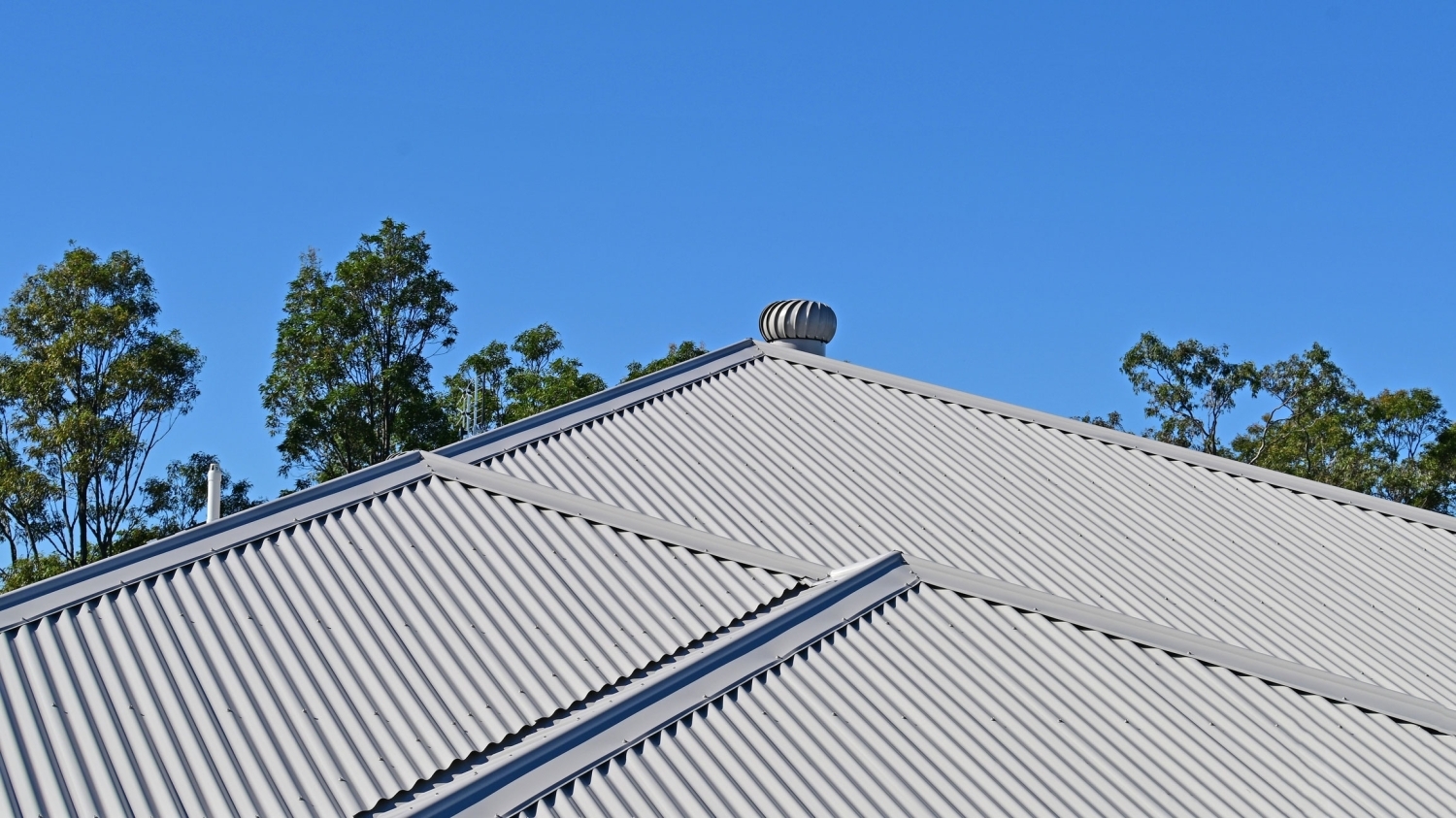 Gray corrugated metal roof with intersecting hips and a turbine vent at the ridge, framed by treetops and clear blue sky.