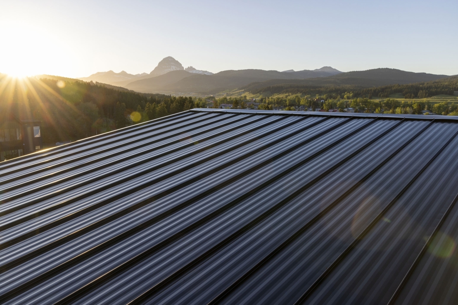 Standing Rib metal roofing in a dark finish, with sunrise light raking across the panels and mountains on the horizon.
