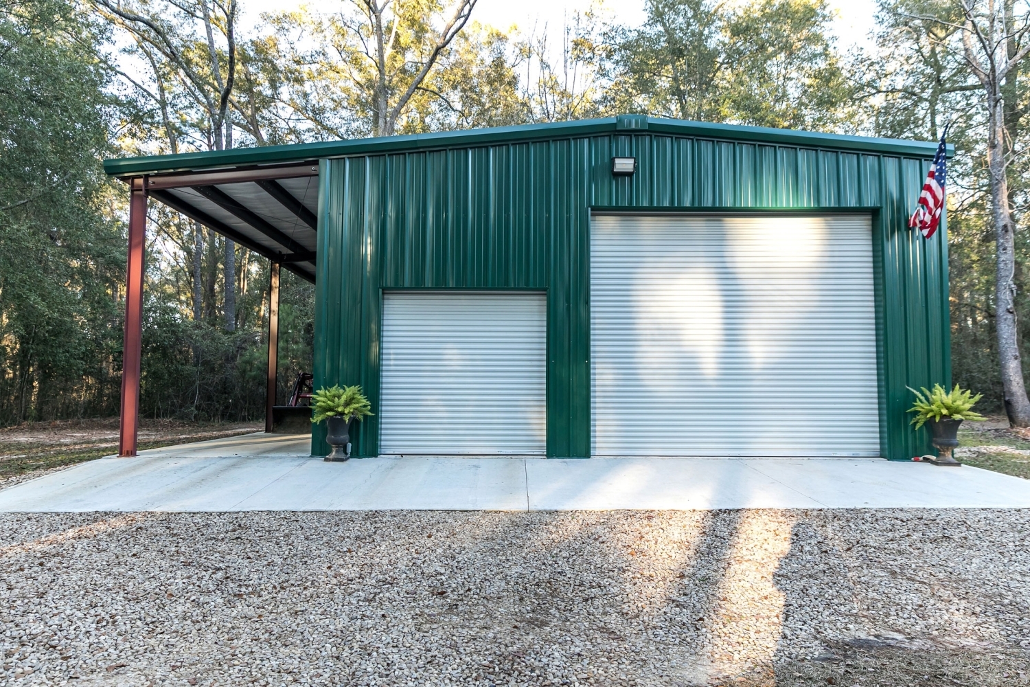 Green metal siding and roofing on a garage with two white roll-up doors, an attached carport bay, and a concrete pad edged in gravel.
