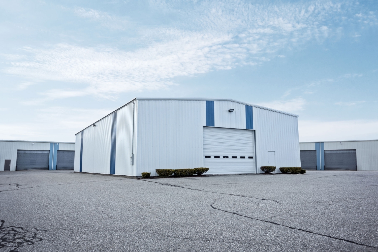 White metal siding with blue accent stripes on a warehouse, with a single overhead door and a wide paved lot in front.