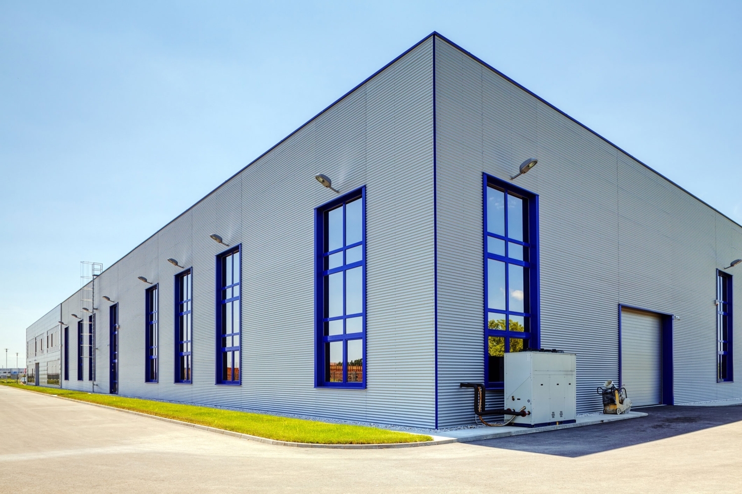 Light gray metal siding on an industrial building with tall blue-framed windows, wall-mounted lights, and a white roll-up door at the corner.
