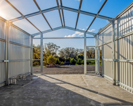 Inside a garage under construction, with a steel frame overhead, cream metal wall panels, and an open end framing the trees beyond.