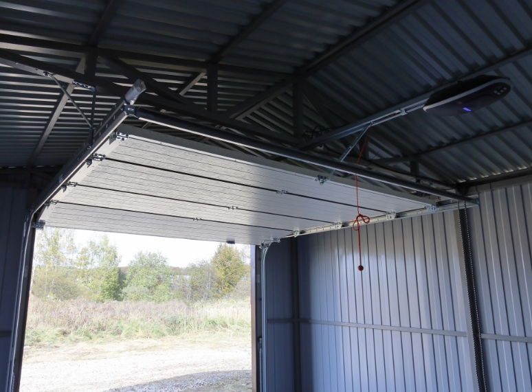Interior of a metal garage with an overhead door raised on its track, exposed roof framing above, and gray metal wall panels along the side.