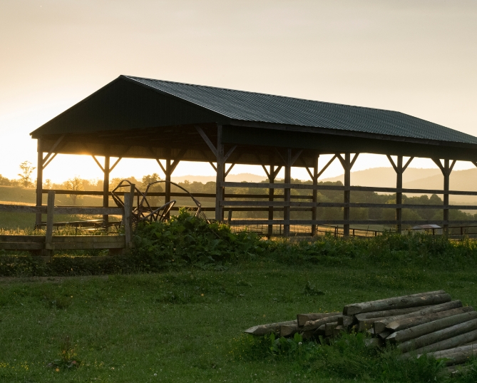 Sunrise behind an open-sided pole shed with a dark metal roof, timber posts, and rolling hills in the distance.