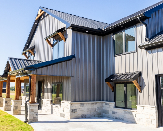 Light gray metal siding and dark metal roofing on a two-story barndominium, with timber porch posts, stone bases, and shed-roof window awnings.