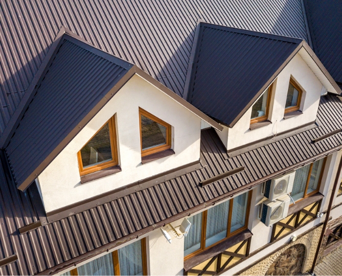 Overhead view of dark corrugated metal roofing across the intersecting gables and dormers of a white stucco house with wood-framed windows.