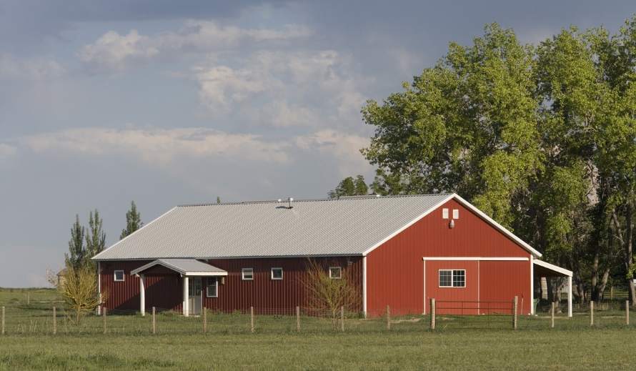 Red metal siding with white trim and a gray metal roof on a farm building, with a covered entry, an attached lean-to, and wire fencing across the pasture in front.