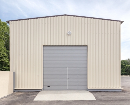 Cream metal siding on a storage building with a dark roof edge, a gray roll-up door, and a concrete ramp leading up to it.