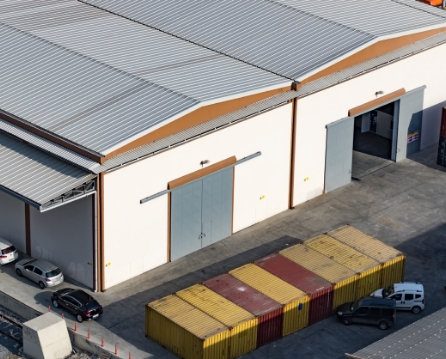 Aerial view of a warehouse with a corrugated metal roof and cream walls, with large sliding doors and shipping containers lined up in the yard.