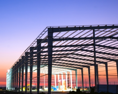 Steel frame of a warehouse under construction at dusk, with columns and roof trusses backlit by an orange and purple sky.
