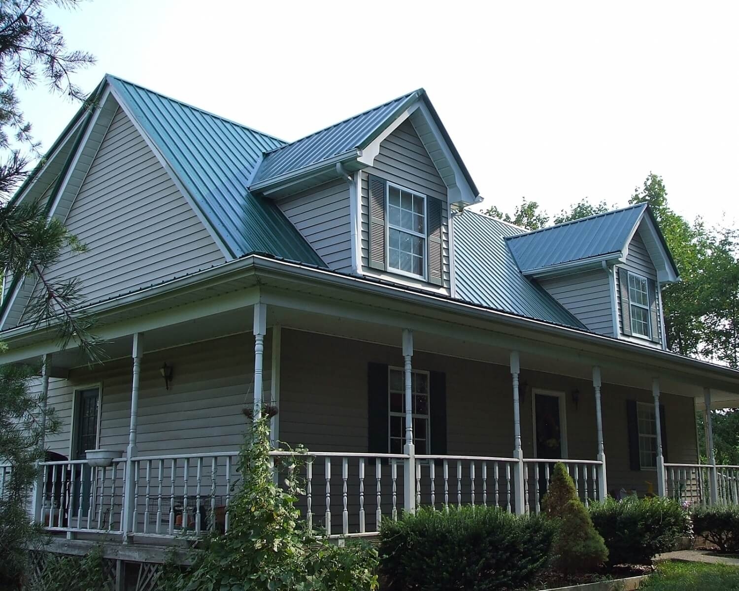 Steep blue metal roofing across the main gable, dormer, and wraparound porch of a two-story home with light gray lap siding.
