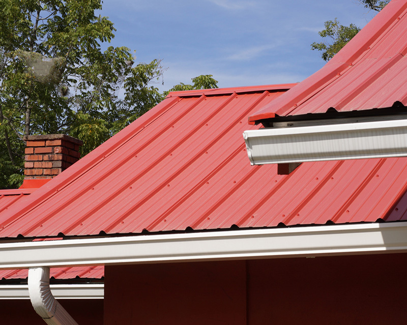 Close view of a red metal roof where two slopes meet at a hip, with a brick chimney, white gutters and fascia, and exposed fasteners along the panel ribs.