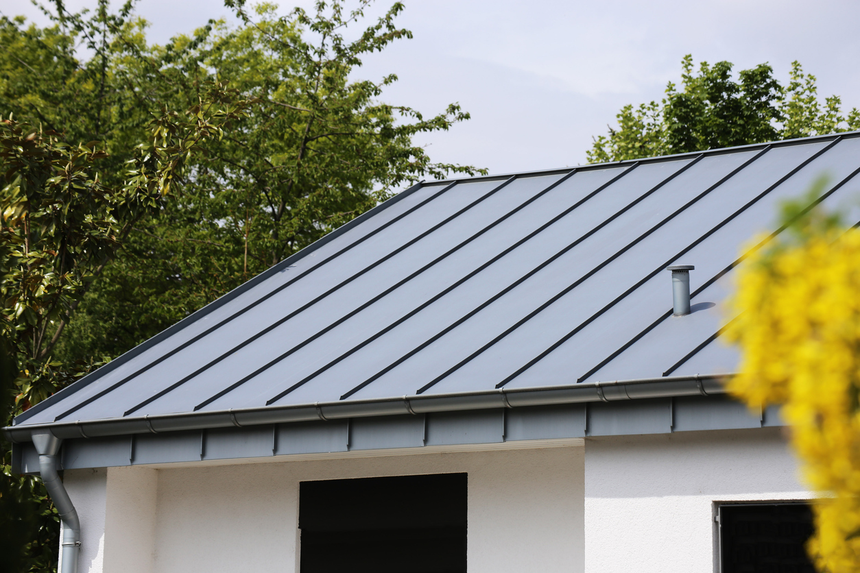 Gray metal roof with dark seams on a white stucco house, with a small vent pipe near the ridge and gutters running the length of the eave.
