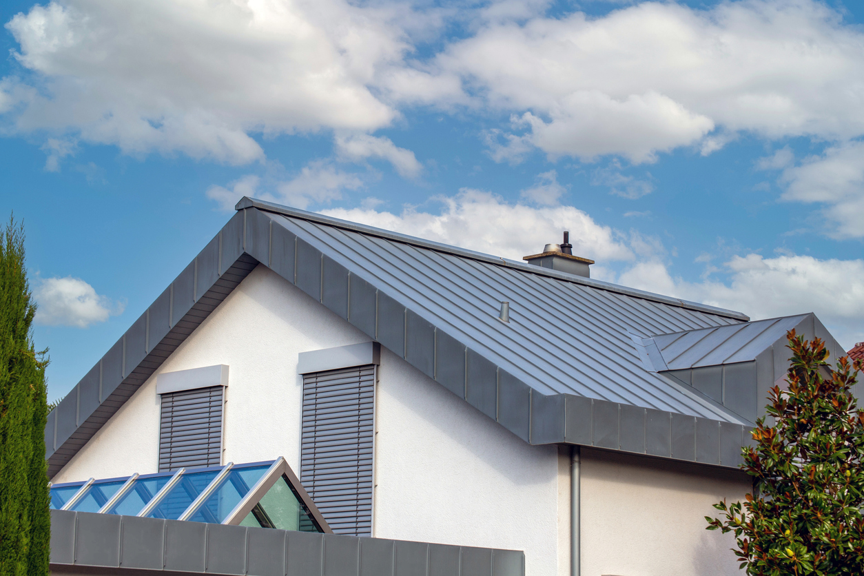 Gray metal roof with wide flashing along the eaves and gable of a white stucco house, with shuttered windows and a glass sunroom at the corner.