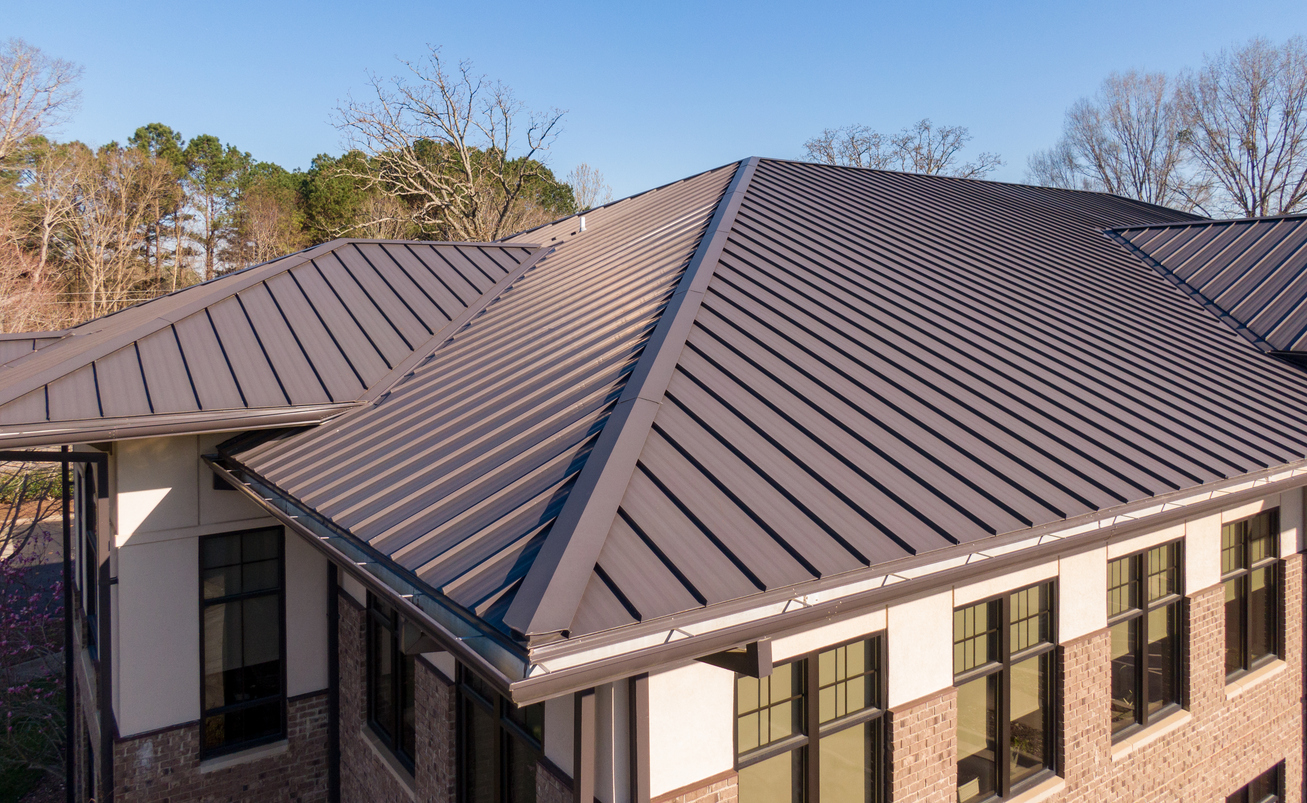 Aerial view of a bronze metal roof over the hips and valleys of a commercial office building, with brick and stucco walls and tall grid windows below.