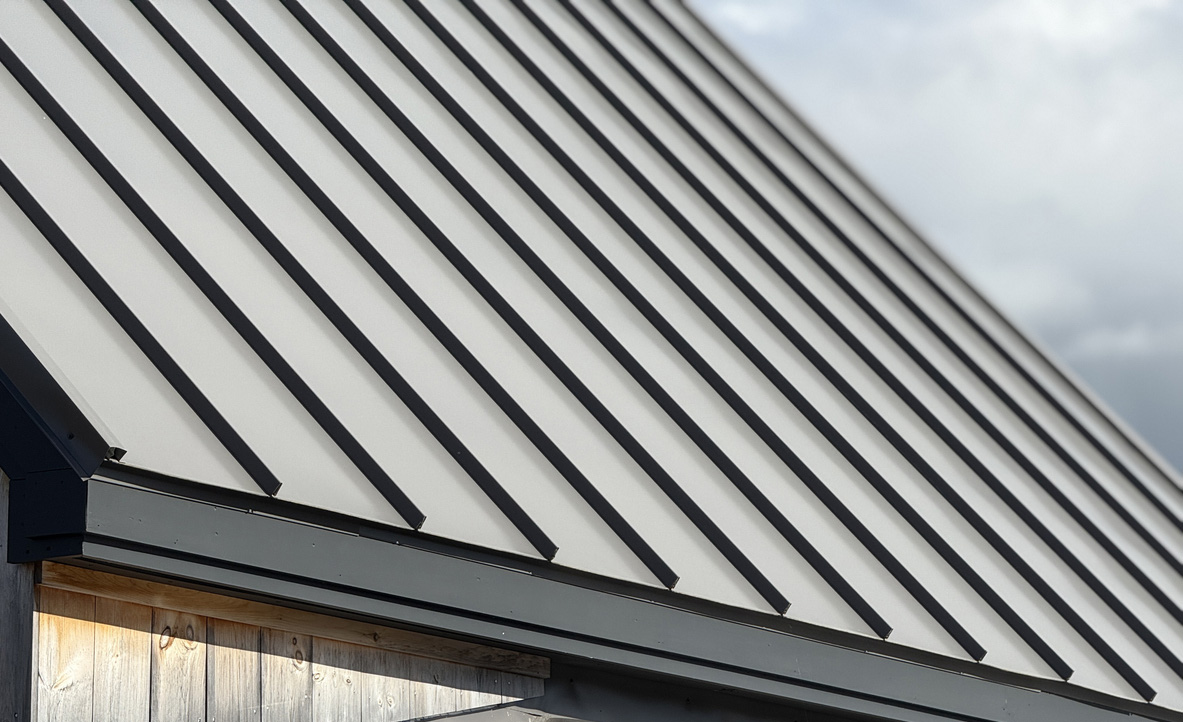 Close view up a cream metal roof with dark seams, ending at a dark eave trim above weathered wood siding.