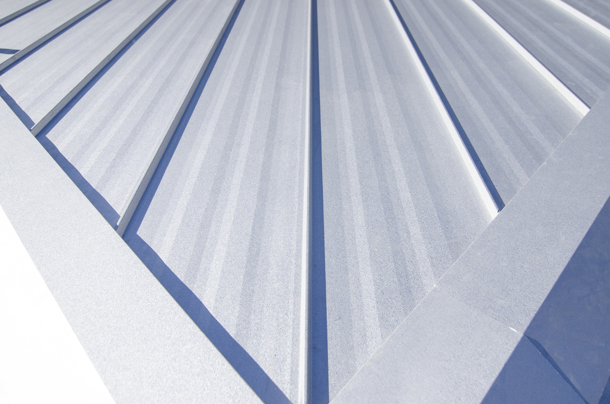 Close view down a white metal roof toward a hip, with sunlight catching the raised seams and shadows falling between them.