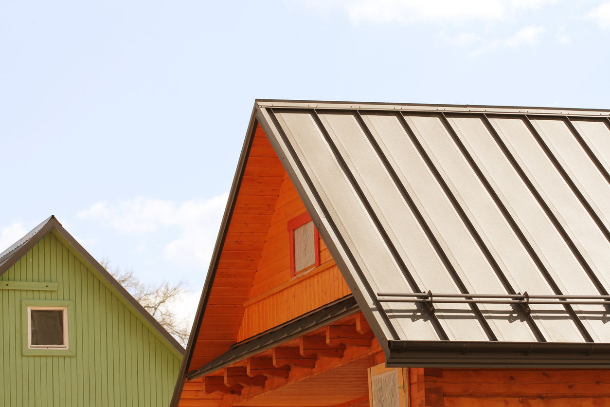 Cream metal roof with brown seams and snow guards on a wood-sided home, with a green-sided gable rising behind it.
