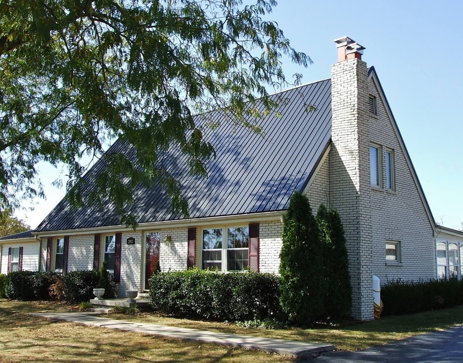 Dark gray metal roof on a white brick home with maroon shutters, a tall brick chimney, and trimmed shrubs along the front walkway.