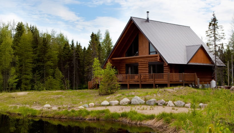 Steep silver metal roof on a log cabin with a wraparound deck, tall gable windows, and evergreens and a pond surrounding it.