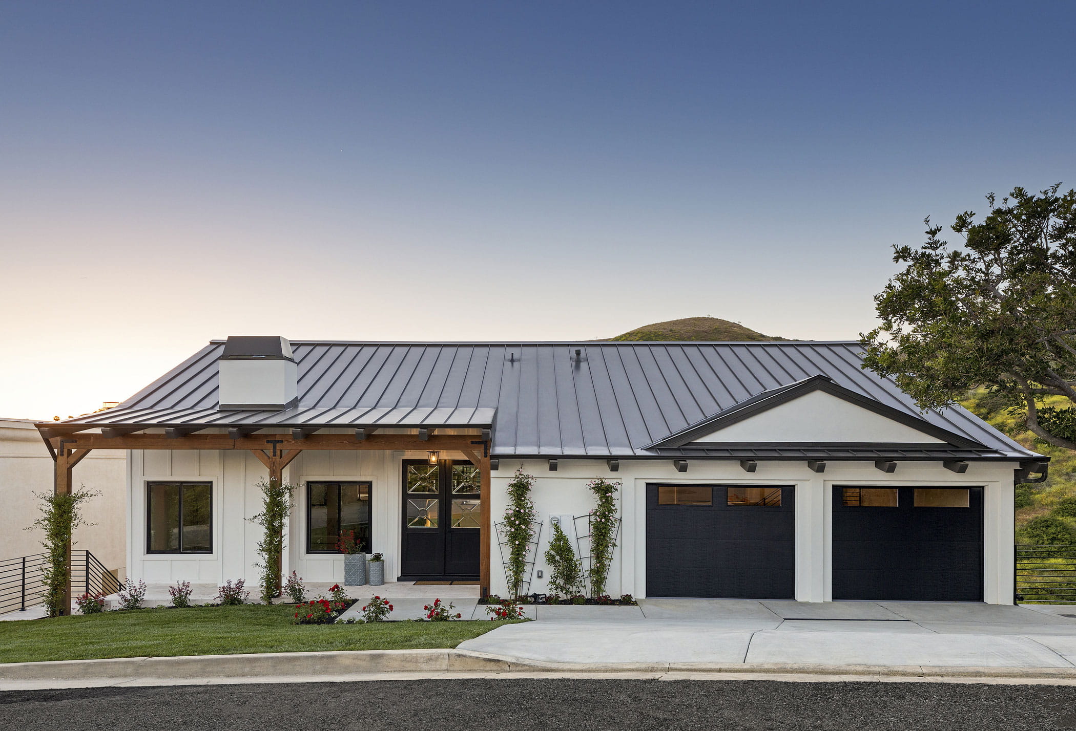Charcoal metal roof on a modern white farmhouse at dusk, with a timber entry trellis, black windows, and two black garage doors.