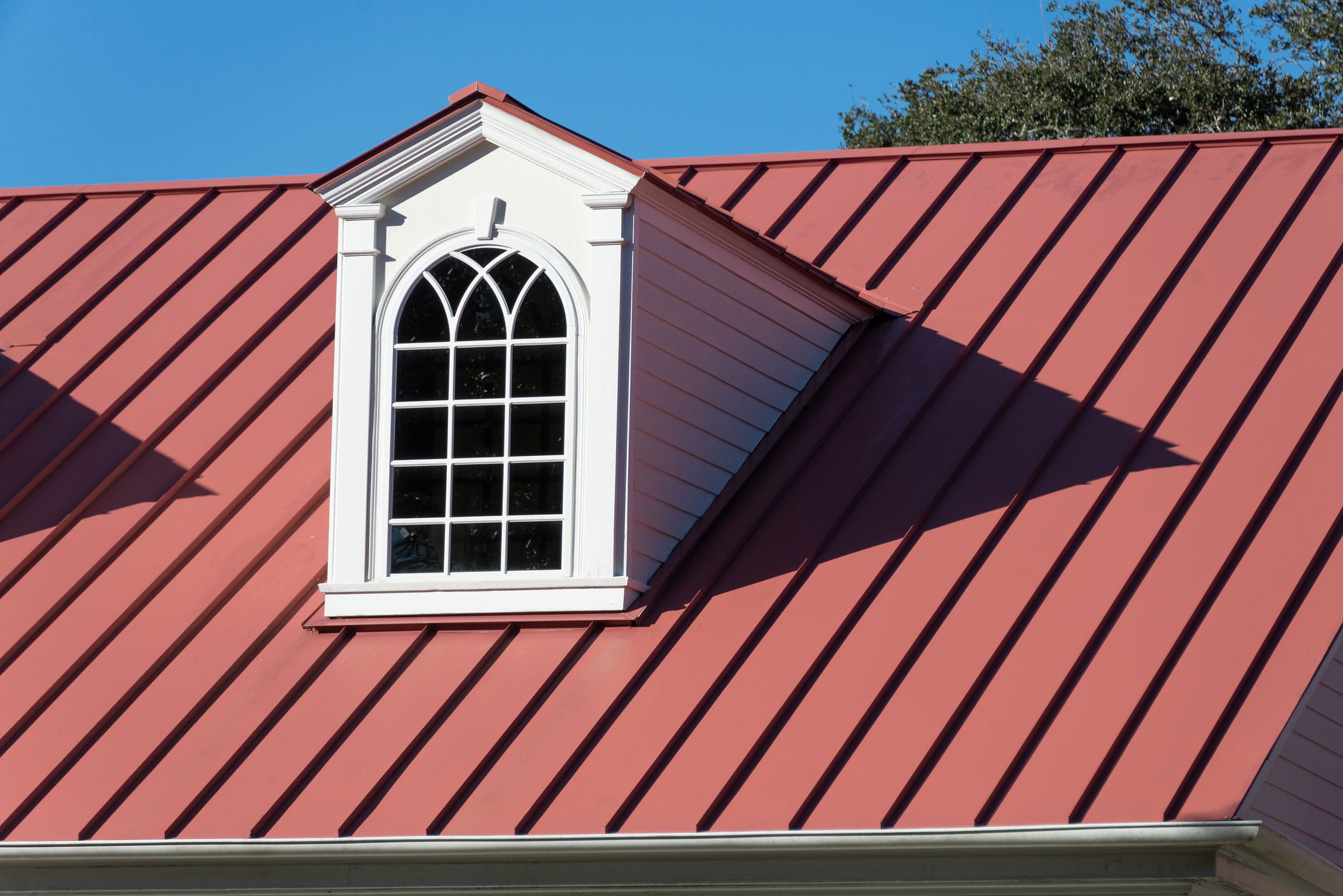 Close view of a red metal roof around a white dormer with an arched, multi-pane window and pedimented trim.