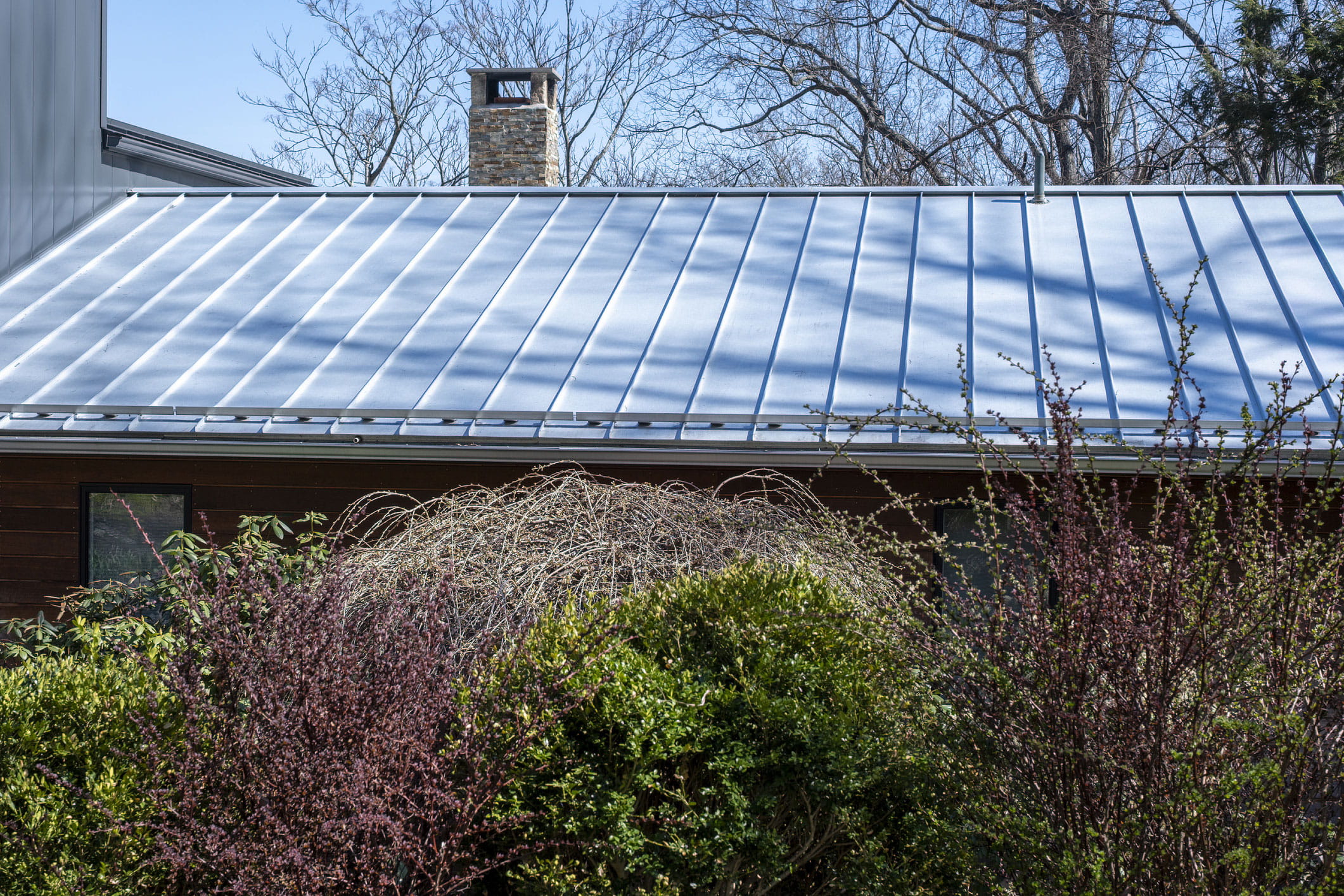 Dark blue metal roof on a house under construction, with fallen leaves collected in the valley beside a dormer and bare framing visible below.