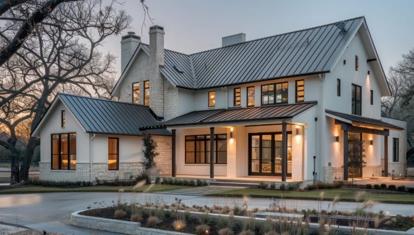 Dark gray metal roofing across the gables and porch of a modern farmhouse at dusk, with white stucco, limestone accents, black windows, and ornamental grasses along the drive.