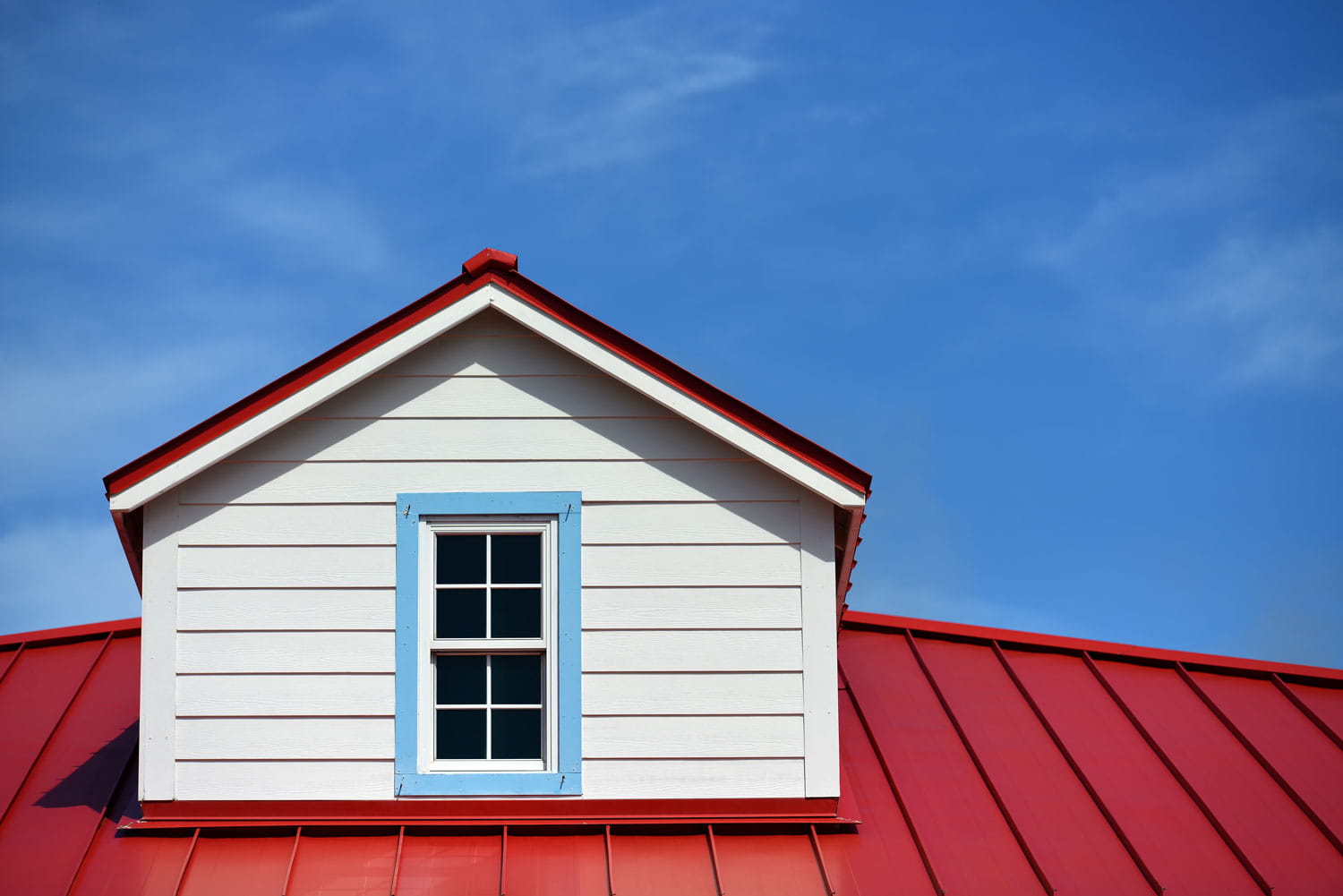 Red metal roof surrounding a white dormer with lap siding, a blue-trimmed window, and blue sky above.
