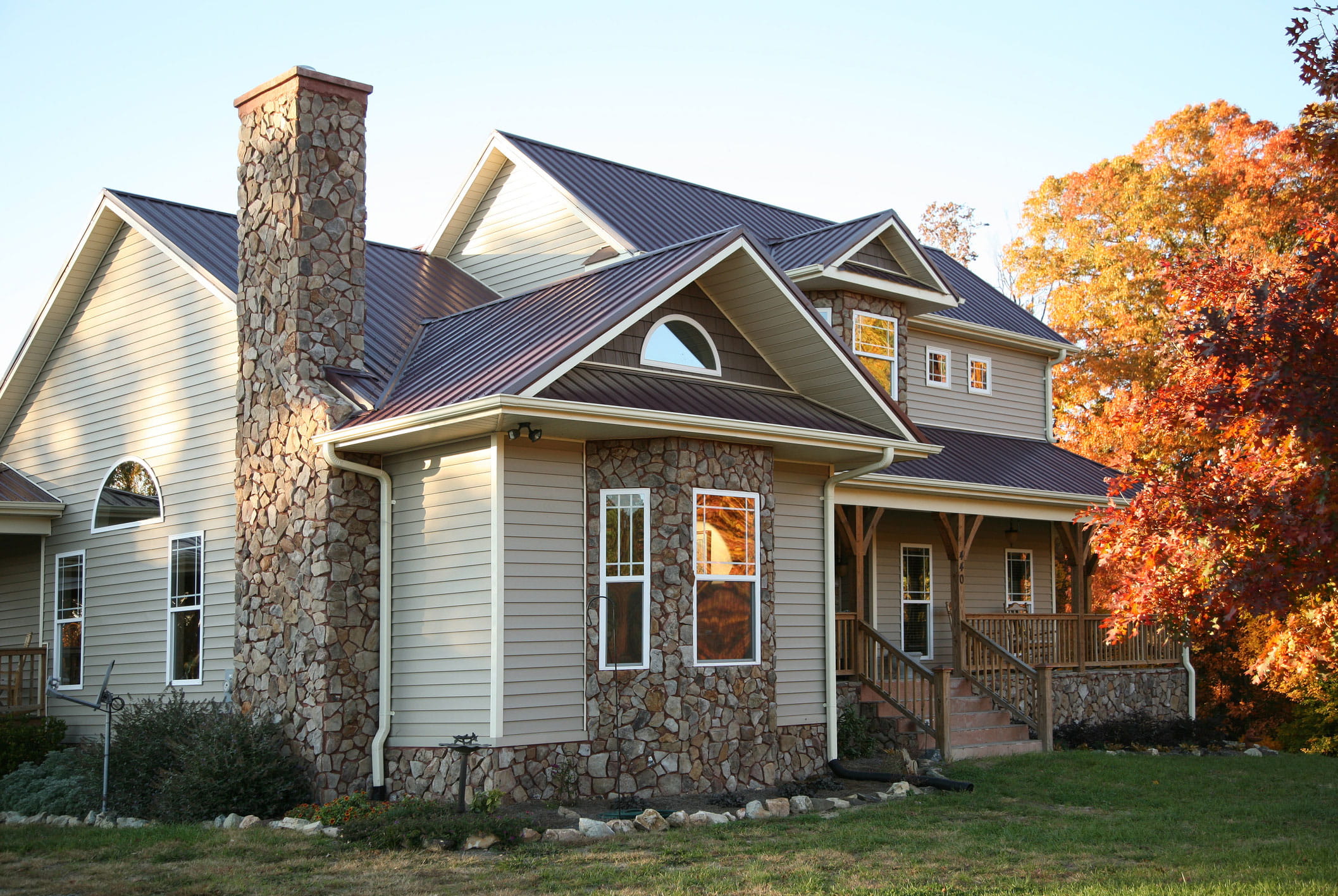 Dark brown metal roofing across the many gables of a two-story home with tan siding, a tall stone chimney, and fall foliage alongside.