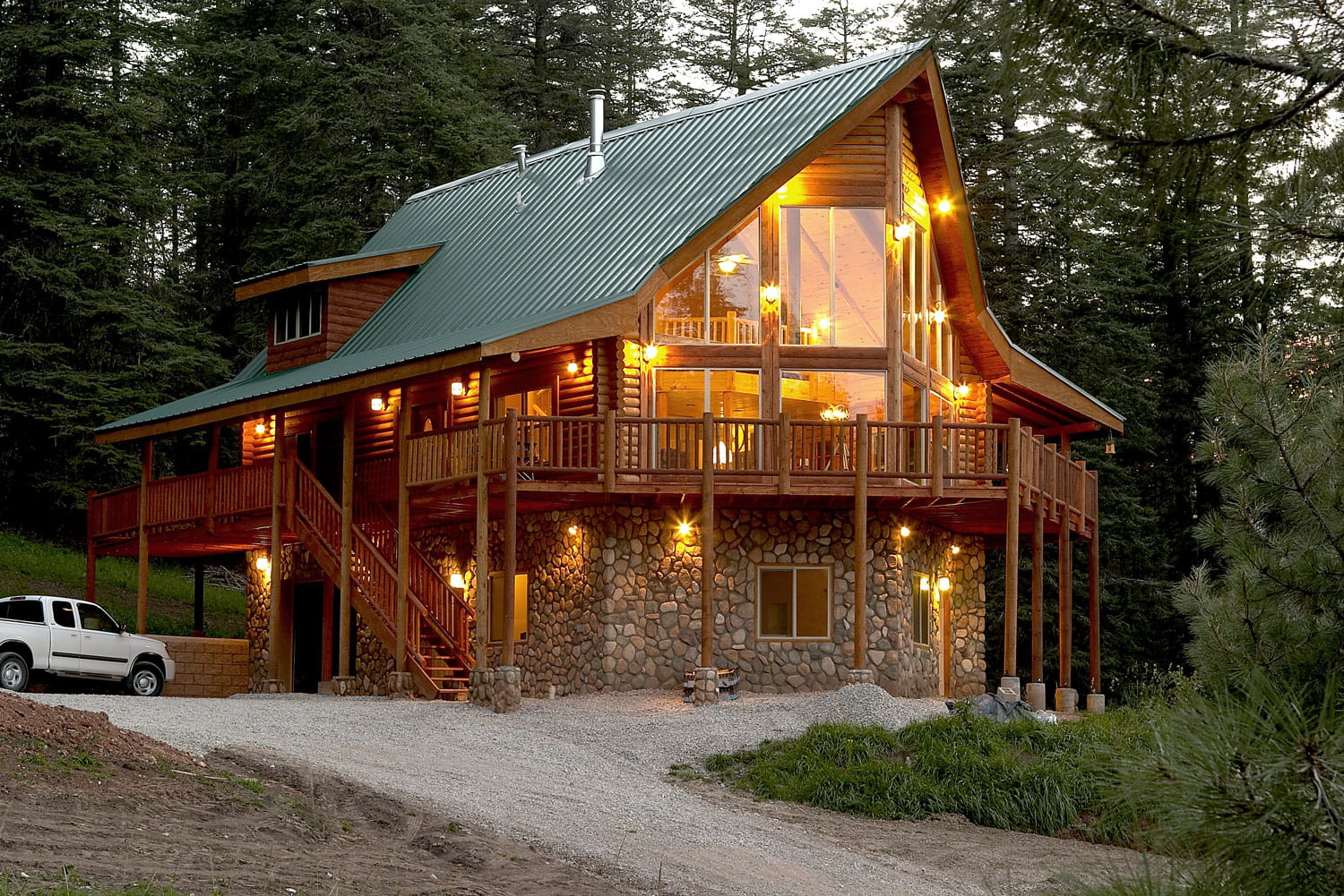 Green metal roof over the steep gable and wraparound porch of a log home at dusk, with river rock foundation walls, tall lit windows, and pines closing in behind.